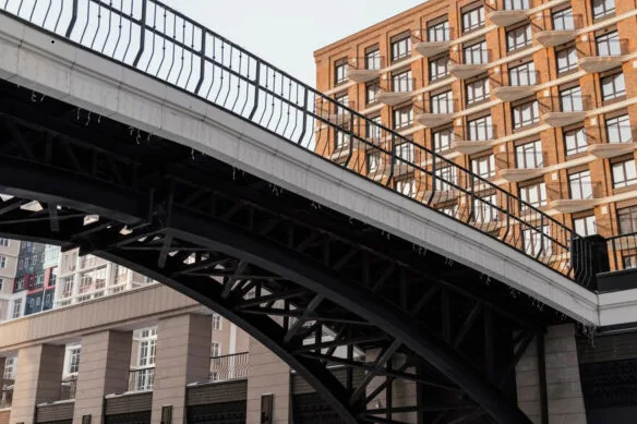 A bridge spans above a street, with a building featuring a repetitive balcony pattern in the background.